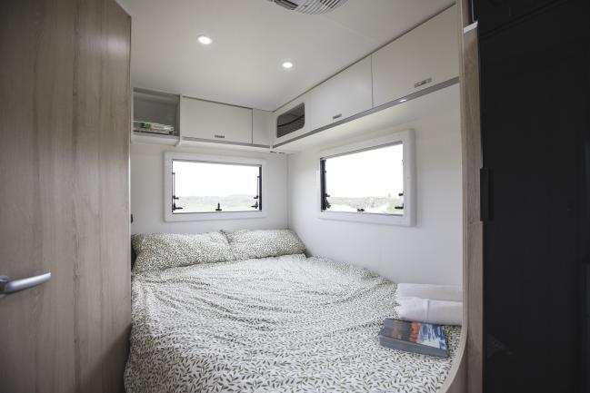 Double bed with leaf-patterned bedding inside a campervan with white overhead cabinets and two windows.