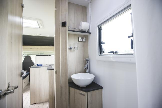 Compact campervan bathroom with a round white basin on a corner cabinet next to a window.