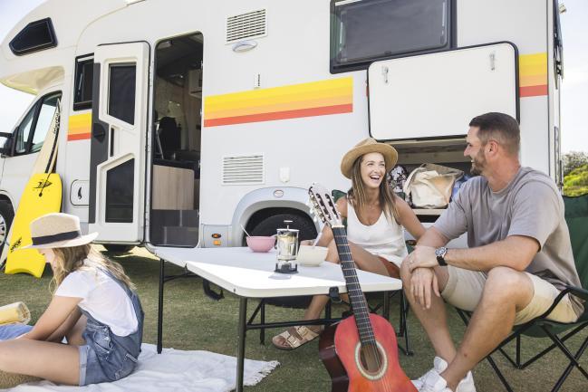 Family relaxing outside a white campervan with camp chairs, a blanket, a guitar, and a surfboard.