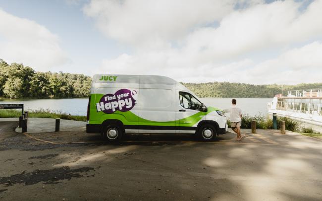 White JUCY campervan with 'Find your Happy' branding parked by a lake, with a person leaning against the front.