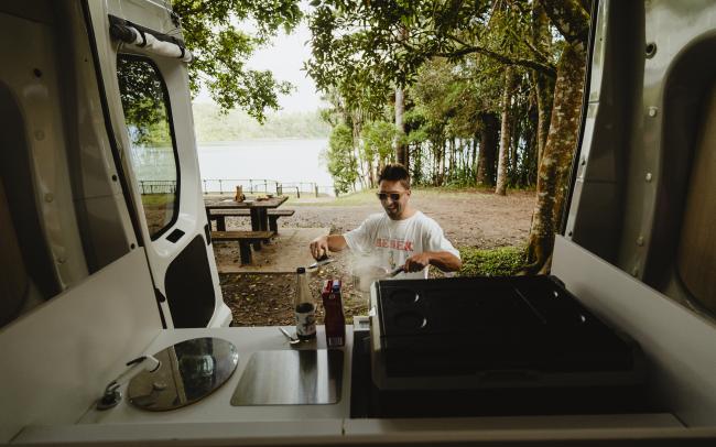 View from a campervan kitchen looking out to a man cooking outdoors, with a lake and forest in the background.