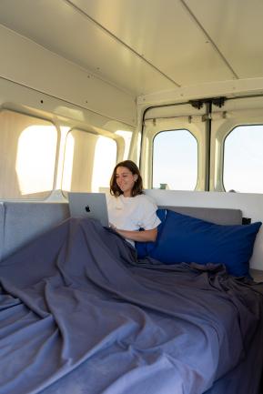 Smiling woman using a laptop in a campervan bed with grey sheets and a blue pillow.