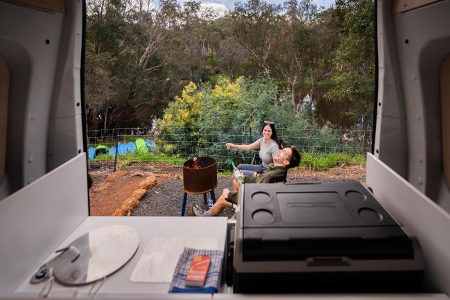 View from inside a campervan looking out at a man and woman laughing by a fire pit near a river.