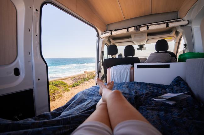 First-person view from a campervan bed, looking past crossed legs out the open door to a sunny ocean coastline.