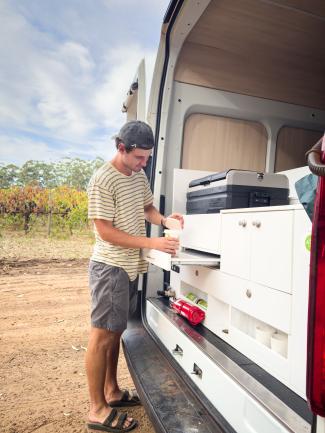 Man prepares food using a slide-out drawer kitchen in the back of a white campervan near a vineyard.