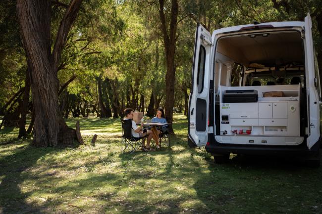 Two people sit at a table next to a white campervan with open rear doors, parked in a grassy clearing under trees.