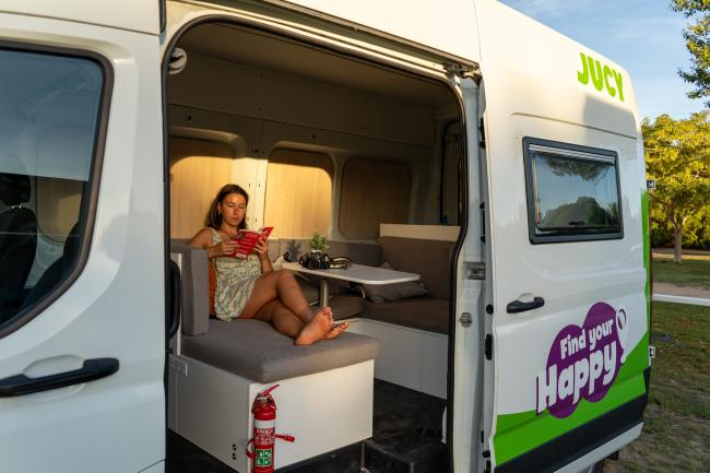 Woman reading a book inside a white Jucy campervan with its side door open, showing the interior seating.
