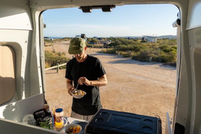 A man in a green cap eats from a bowl outside the open back of a campervan parked in a coastal landscape.