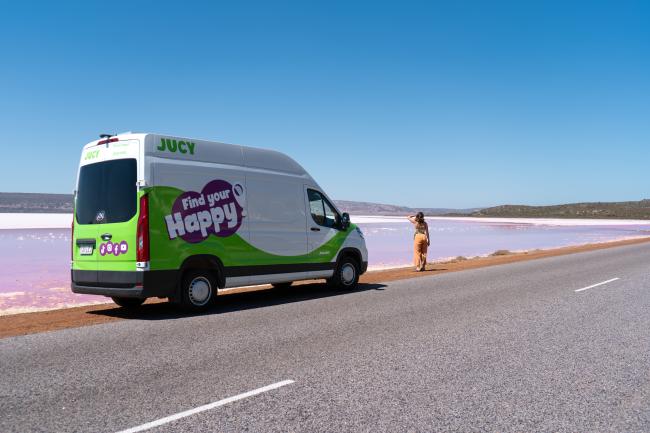 White and green JUCY campervan parked beside a pink lake under a blue sky, with a person standing on the shore.