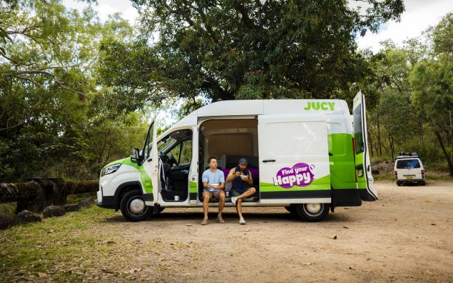 Two men sit in the open side door of a green and white JUCY campervan parked in a clearing surrounded by trees.