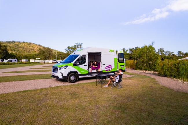 White and green JUCY campervan parked at a campsite, with one person resting inside and another sitting outdoors.