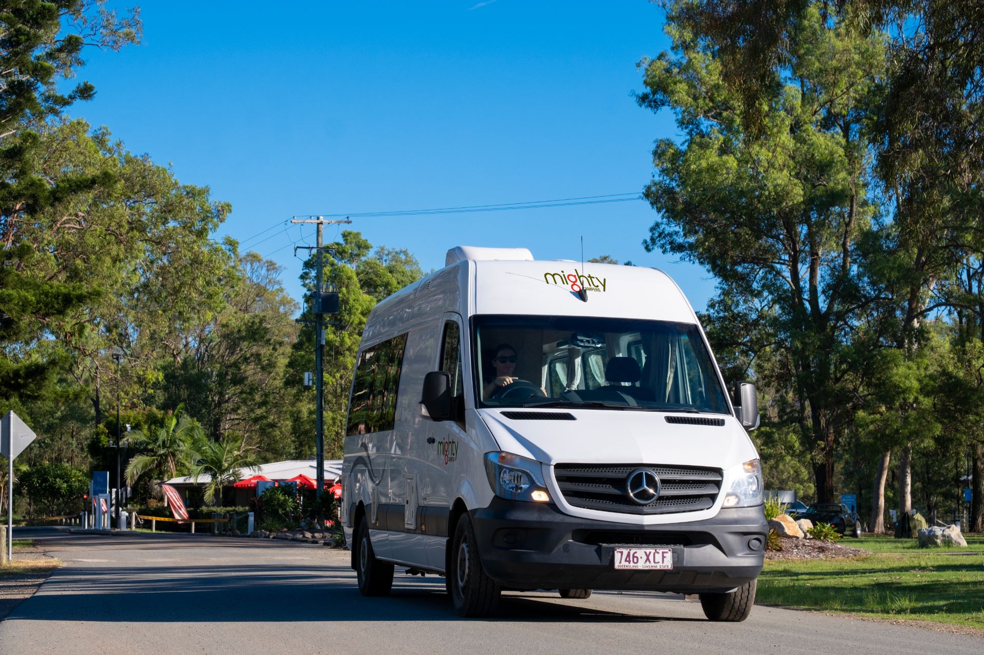 White Mighty Campers Mercedes campervan driving on a tree-lined road under a clear blue sky.