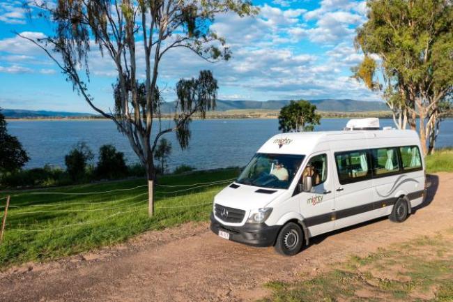 White Mighty campervan parked on a dirt track next to a grassy area overlooking a lake.