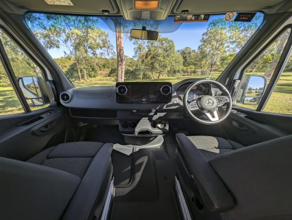 Right-hand drive Mercedes campervan cab interior with grey seats, steering wheel, and dashboard looking out at trees.