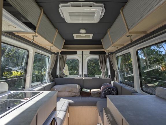 Campervan interior featuring grey U-shaped seating, kitchen counters, overhead storage, and large side windows.