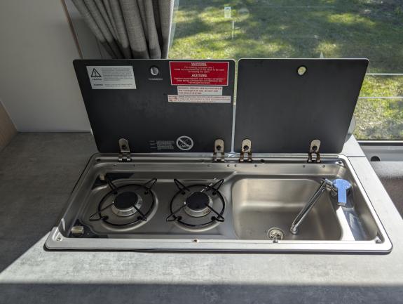 Campervan stainless steel unit with a two-burner gas stove, sink, fold-down tap, and raised black glass lids.