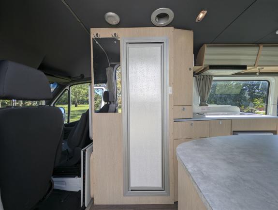 Interior of a campervan showing front seats, a light timber cabinet with a frosted door, and a kitchen counter.