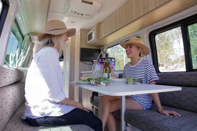 Two women wearing hats sit opposite each other, chatting over a meal inside a campervan.