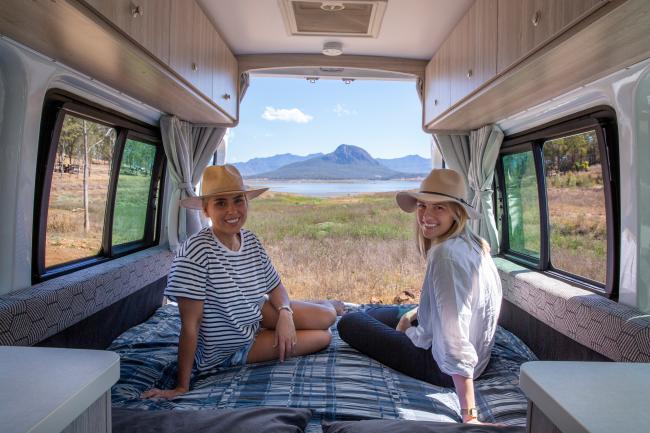 Two women wearing sun hats sit inside a campervan with open back doors showing a lake and mountain landscape.