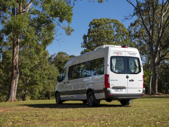 White Mighty Mercedes-Benz campervan parked on grass, surrounded by tall trees under a blue sky.