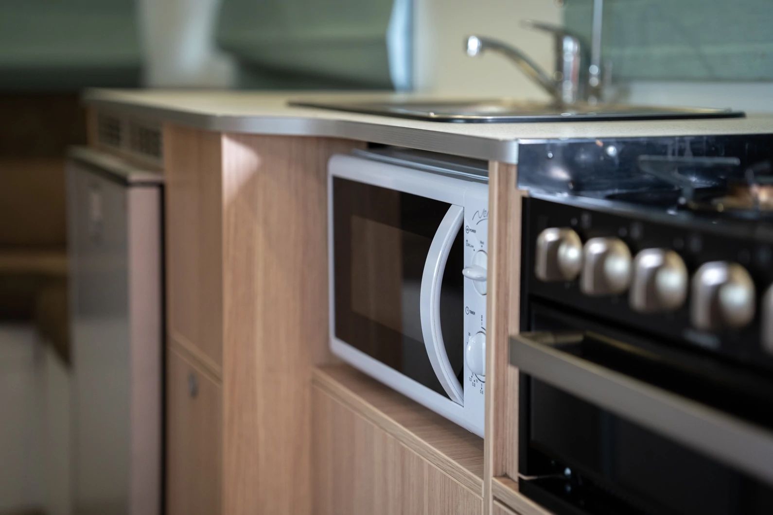 White microwave built into light timber cabinetry next to a black stove in a compact campervan kitchen.