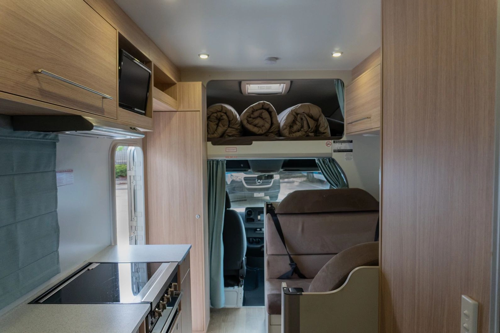Compact campervan interior featuring a kitchenette, dinette seat, light timber-look cabinets, and an overcab bed.