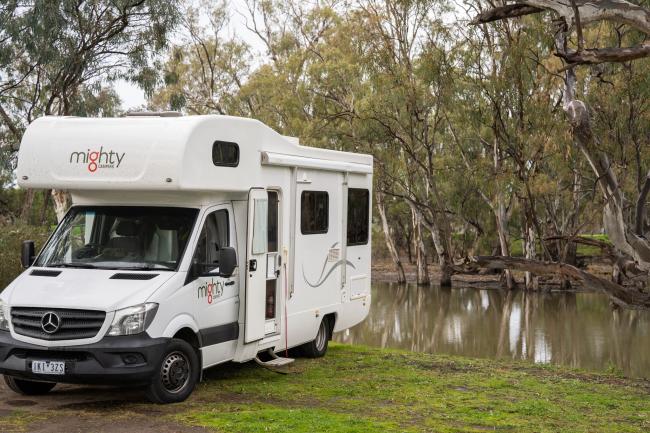 White Mighty campervan parked on a grassy bank beside a river lined with gum trees.