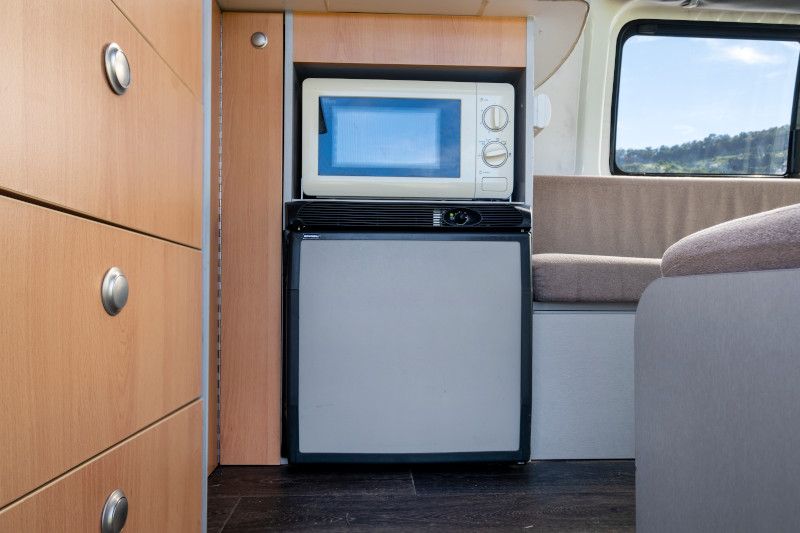 Built-in microwave and compact refrigerator next to light timber drawers inside a campervan.