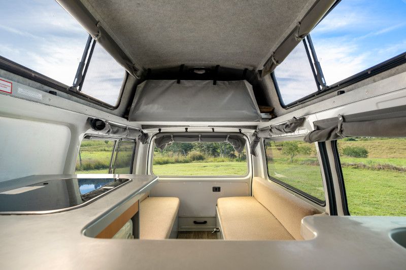 Interior of a campervan featuring twin beige bench seats, a kitchen counter, and windows overlooking a grassy field.