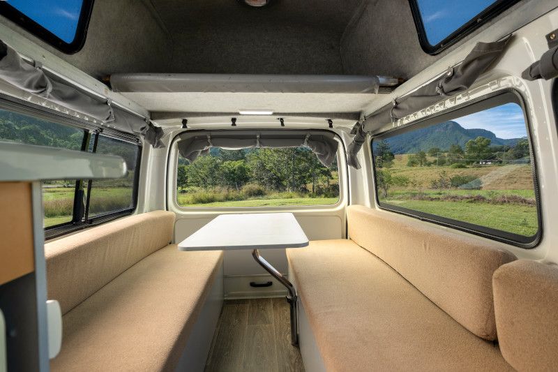 Campervan interior with parallel tan bench seats, a central white table, and large windows showing grassy hills outside.