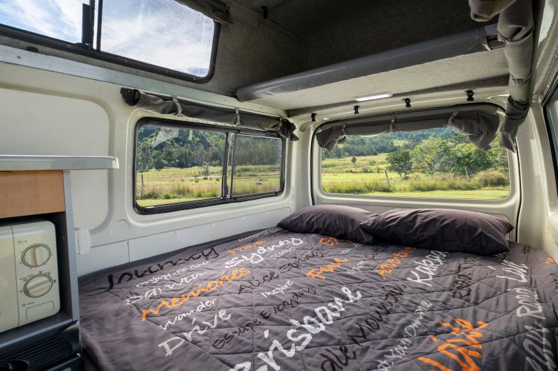 Campervan interior with a double bed made with a grey text-print quilt, looking out windows onto green fields.