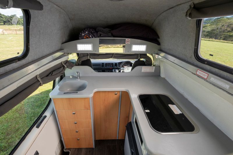 Campervan interior showing a grey countertop with a sink and stove, timber drawers, and overhead storage.