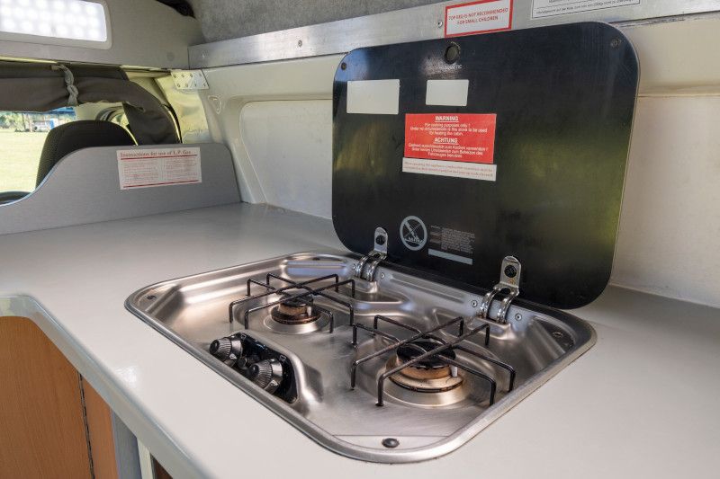 Two-burner gas stove with an open black lid built into a light grey campervan kitchen benchtop.