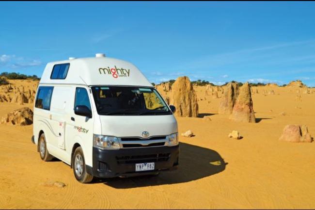 White Mighty high-top campervan parked in a sandy desert with limestone pillars under a blue sky.