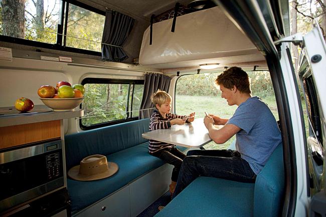 A man and child play cards inside a campervan, sitting at a table on blue bench seats.