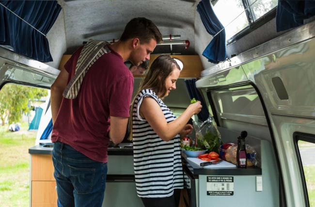 A man and woman prepare salad together inside a compact campervan kitchen.