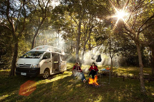 Two people camp beside a white campervan in a forest clearing, sitting by a campfire under streaming sunbeams.