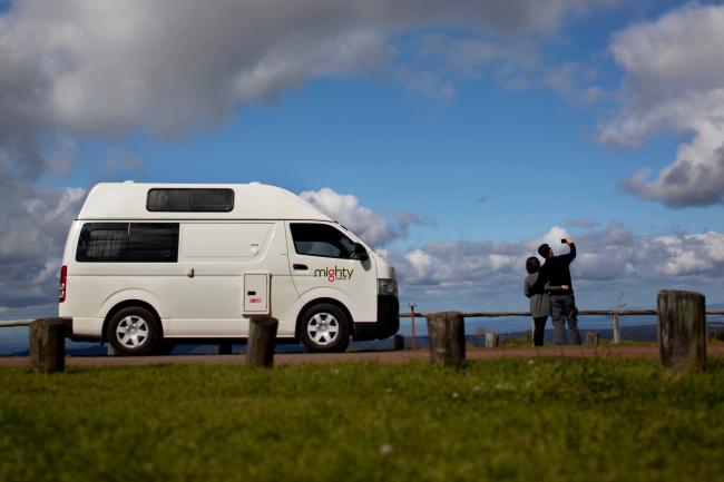 White Mighty campervan parked at a grassy lookout with two people taking a selfie in the background.