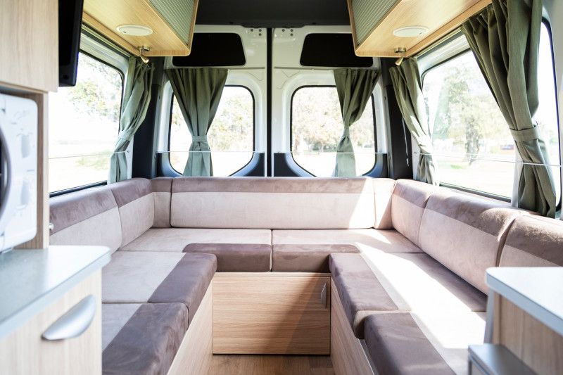 Campervan interior featuring a U-shaped beige lounge, light timber cabinets, and tied-back sage green curtains.
