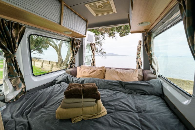 Campervan interior featuring a double bed with grey bedding and folded towels, looking out to a coastal sea view.