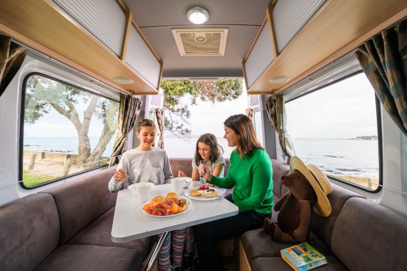 A woman and two girls eat breakfast at a table inside a campervan with coastal views through the windows.