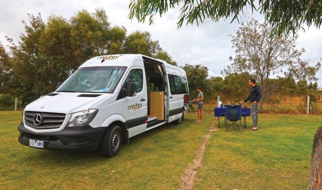 White Mighty campervan parked on grass with two people preparing a meal at an outdoor table.
