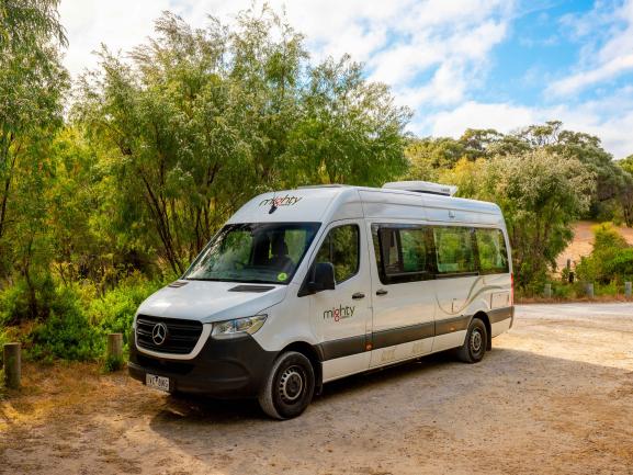 White Mighty Mercedes-Benz campervan parked on a gravel road surrounded by green trees.