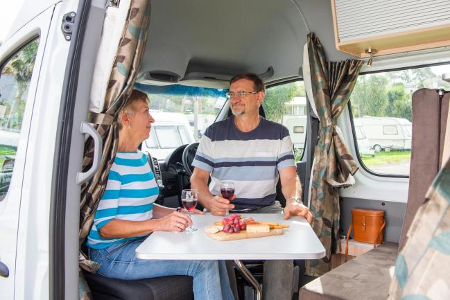A couple sits at a table inside a campervan, enjoying wine and a platter of fruit and cheese.