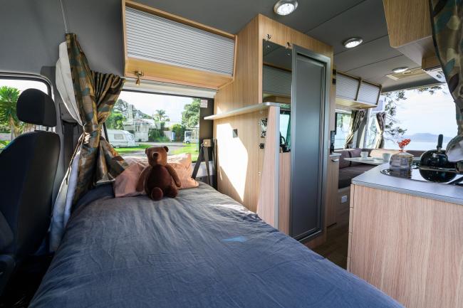 Inside a campervan showing a bed with a teddy bear, light timber cabinets, and a small kitchen.
