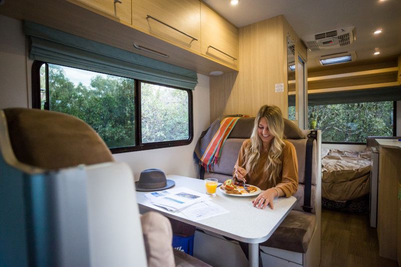 Woman eats a meal at a table inside a campervan next to a window overlooking green trees.
