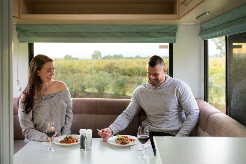 A man and woman eating a meal with wine inside a campervan booth, smiling next to a large window.