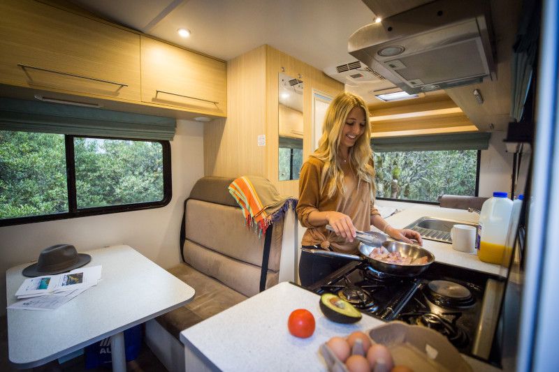 Woman cooking on a gas stove inside a modern campervan kitchen, with fresh ingredients on the counter.