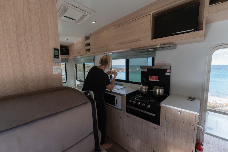 Woman standing in a campervan kitchen with timber cabinetry, looking out the window at the ocean.