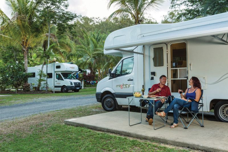 Couple relaxing outside a white Mighty campervan at a campsite, with another campervan parked in the background.